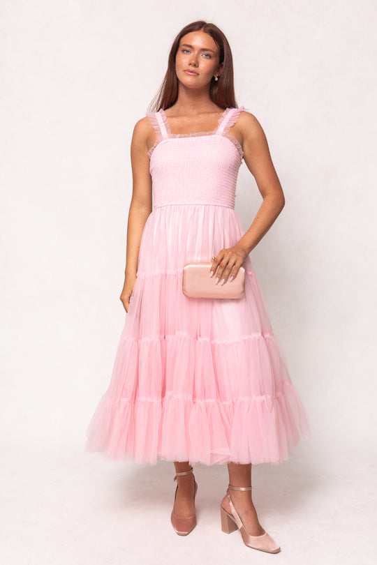 A woman wears the Ivy City Co Destiny Dress in Pink, a sleeveless tulle maternity style, and holds a matching pink clutch. She completes the look with blush heels and poses against a neutral background.