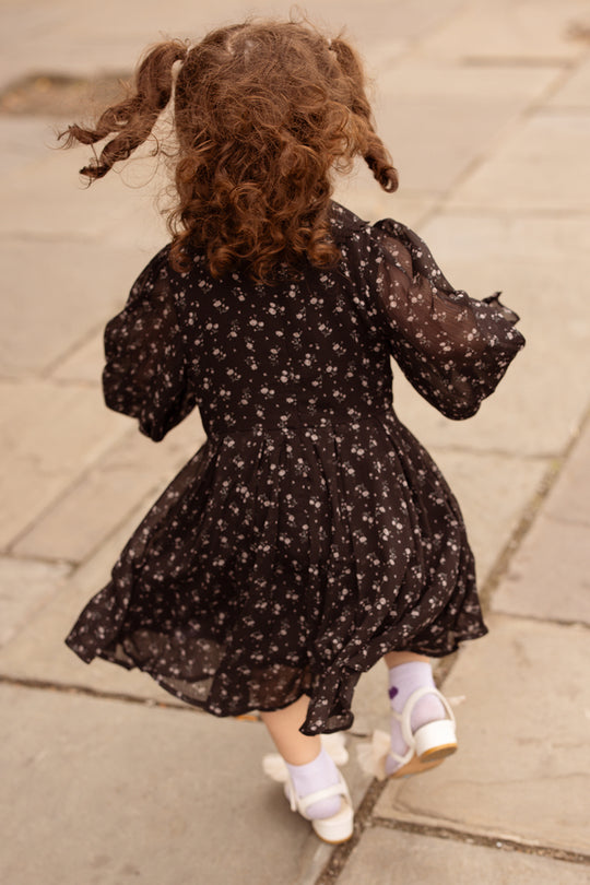 A young child with curly hair in pigtails, wearing the Ivy City Co Mini Sophia Long Sleeve Dress in Noir Ditsy Chiffon and white shoes, is walking away on a stone pavement.