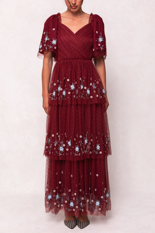 A woman wearing the Mabel Dress in Burgundy by Ivy City Co—a maxi tulle dress featuring short sleeves, light blue and white floral embroidery, a V-neckline, and polka dot detailing—stands against a plain background.