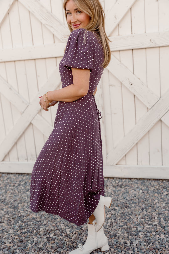 A woman in the Ivy City Co Diane Dress in Aubergine Ditsy Dot—a nursing-friendly, tea-length piece—stands sideways on gravel before a white wooden fence, smiling subtly at the camera in her white ankle boots.