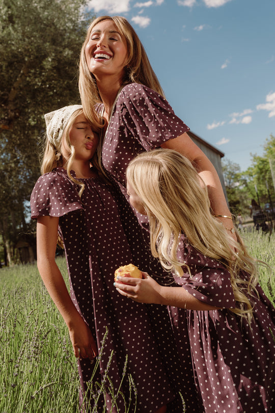 A smiling woman and two girls wear matching Ivy City Co Diane Dresses in Aubergine Ditsy Dot as they stand together in a grassy field. The tea-length, nursing-friendly dresses are perfect for family outings. One girl holds a pastry, and the other wears a light-colored headscarf.