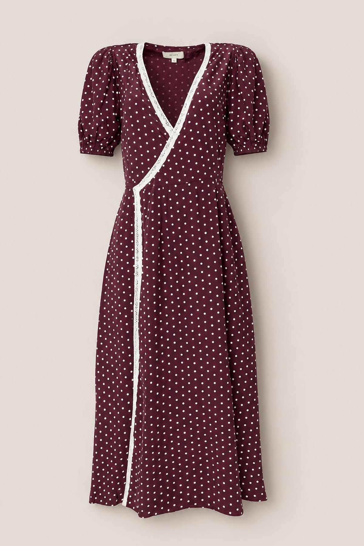 The Diane Dress in Aubergine Ditsy Dot by Ivy City Co features a rich burgundy hue adorned with white polka dots, short puffed sleeves, and delicate white lace trimming the neckline and wrap edge. Its flattering tea-length silhouette exudes timeless elegance—beautifully showcased against a plain light background.