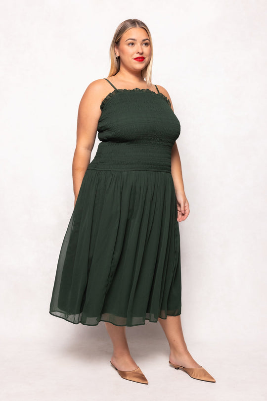 A woman stands against a plain white background wearing the Ivy City Co Kimberly Dress in Green—a dark green, sleeveless midi dress with thin straps and a smocked bodice, paired with tan flats. This maternity-friendly dress effortlessly combines comfort and style.