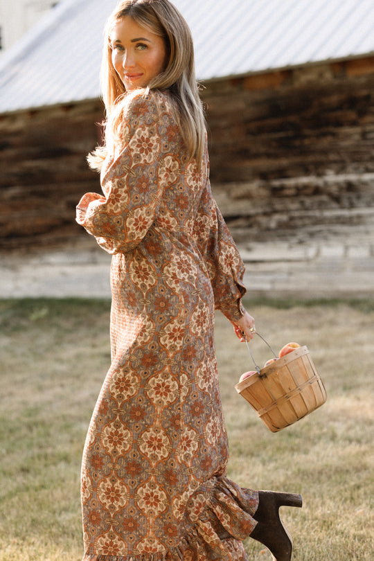 A woman wearing the Ivy City Co Draper Dress in Golden Hour Tesserae, featuring elegant balloon sleeves and a flowing maxi silhouette, walks on grass with a basket of apples in hand, glancing back as a rustic wooden building stands in the background.