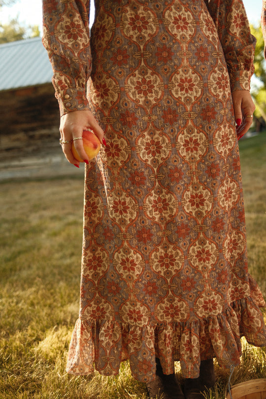 A person wearing the Draper Dress in Golden Hour Tesserae by Ivy City Co, featuring balloon sleeves and a long, patterned design, holds a peach while standing on grass with a rustic wooden building and trees in the background. Only their torso and legs are visible.