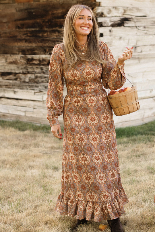 A woman wearing the Ivy City Co Draper Dress in Golden Hour Tesserae, featuring balloon sleeves, stands smiling on dry grass in front of a rustic wooden wall while holding a small basket of apples.