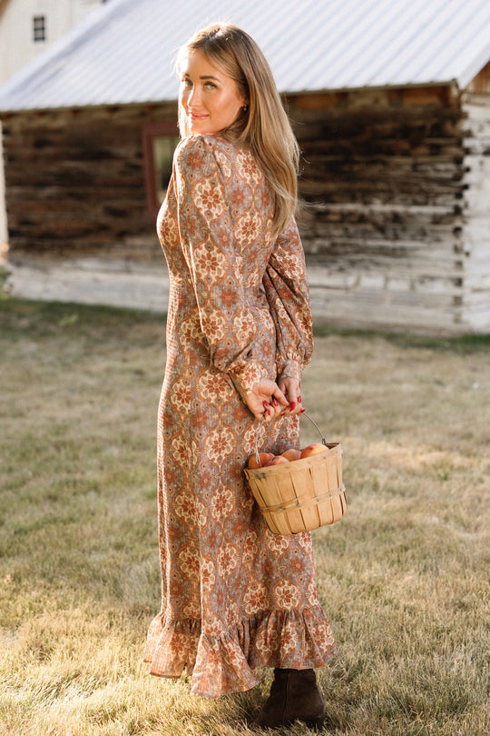 A woman wearing the Ivy City Co Draper Dress in Golden Hour Tesserae, featuring elegant balloon sleeves and a long, floral design, stands on the grass holding a basket of apples and looking back over her shoulder. An old wooden building can be seen in the background.