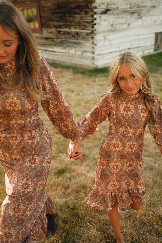 A woman and a young girl wearing matching Ivy City Co Draper Dresses in Golden Hour Tesserae, featuring charming balloon sleeves, hold hands as they walk on the grass near a rustic wooden building.