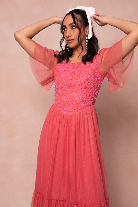 A woman wears the Dulce Dress in Pink by Ivy City Co, featuring short flutter sleeves and tulle fabric, as she ties a large white bow in her hair. She stands against a beige background with statement pearl earrings, gazing sideways.