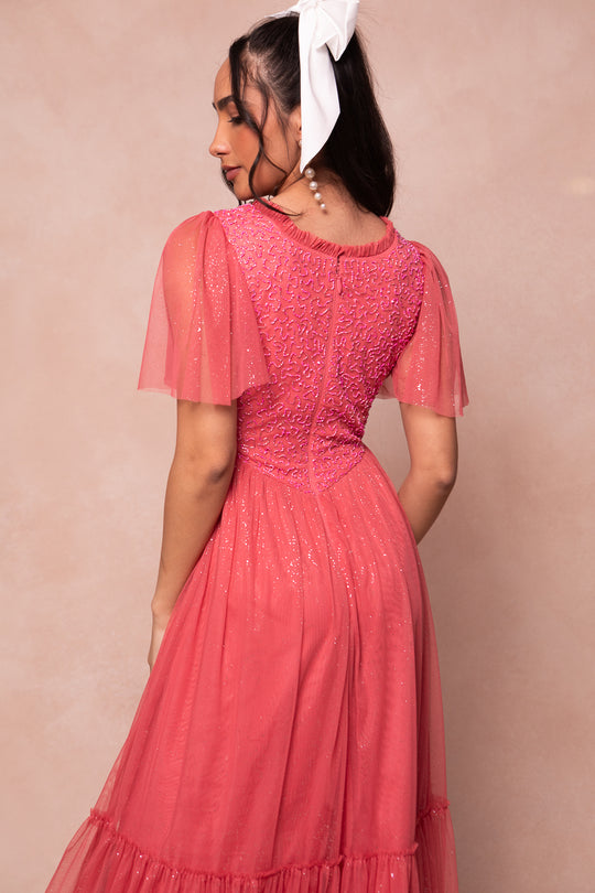A woman with long dark hair tied with a large white bow is seen from the back wearing the sparkly Dulce Dress in Pink by Ivy City Co, featuring short sheer sleeves and a textured bodice, set against a soft beige background.