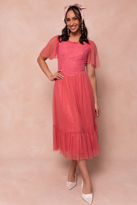 A woman smiles in the Dulce Dress in Pink by Ivy City Co, featuring glitter details, flutter sleeves, and a ruffled hem. She pairs it with a matching headscarf and white pointed shoes, standing before a light pink backdrop.