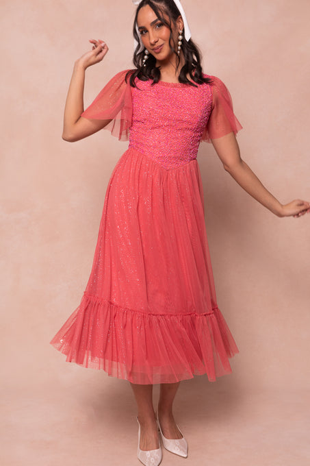 A woman poses against a light beige backdrop wearing the Ivy City Co Dulce Dress in Pink, featuring sparkly fabric, short flutter sleeves, and a ruffled hem. She accessorizes with a white headband and pointed-toe shoes.