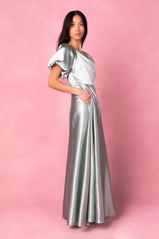 Dressed in the Ivy City Co Tessie Dress in Sage, a woman stands against a pink background and looks at the camera, her left hand casually tucked into the dress pocket—a stylish, modest choice that blends elegance with comfort and would make an excellent bridesmaid dress.