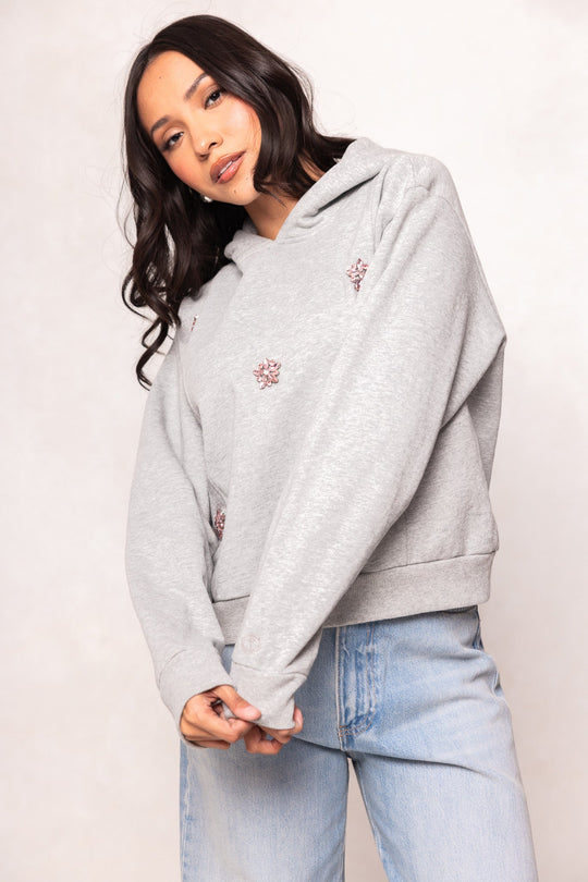 A woman with long dark hair wears the Ivy City Co Skylar Hoodie in heather gray, featuring small embroidered designs, paired with light blue jeans. She stands against a pale background, gazing slightly downward.