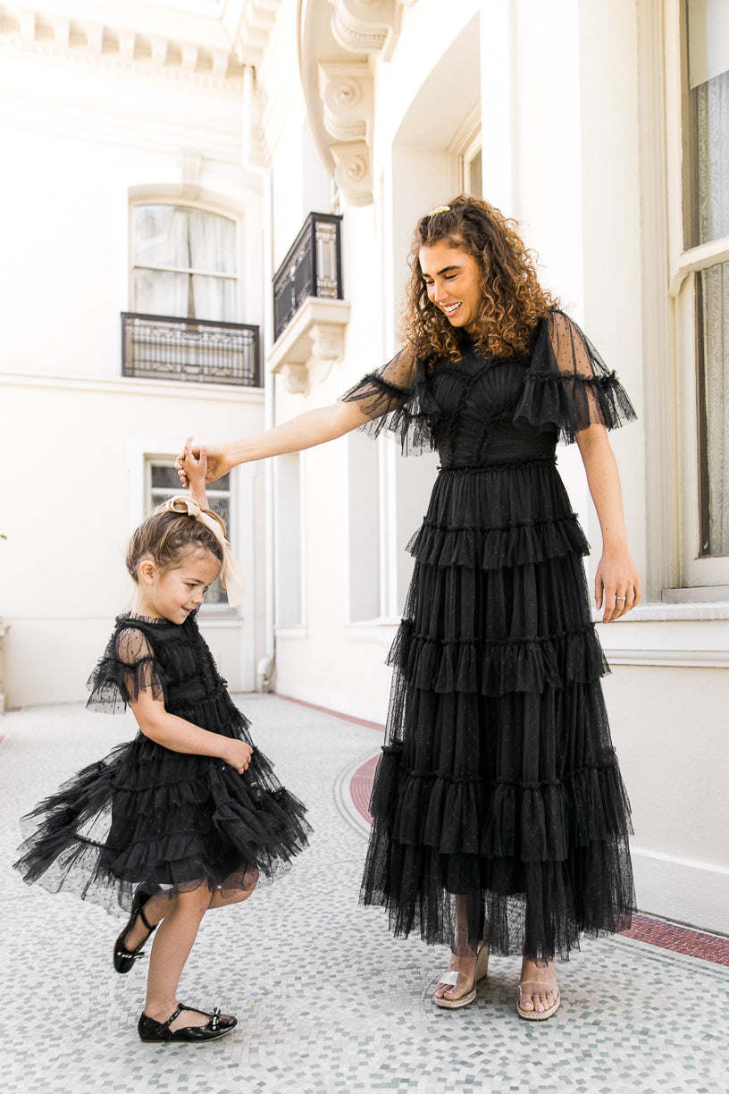 A woman and a young girl, both wearing the Ivy City Co Mini Whimsical Dress in Black with ruffled details and layered tulle skirts, dance together outside a white building. The woman holds the girl’s hand, smiling, while the girl twirls with her head lowered.