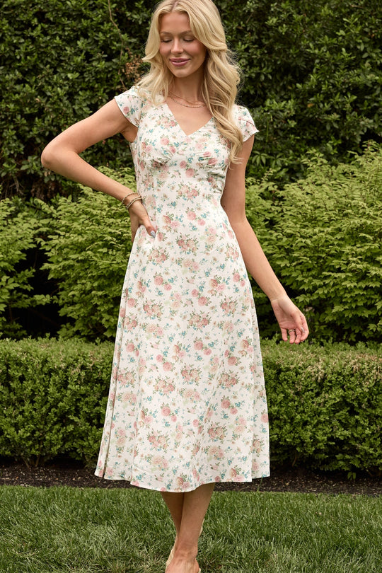 A woman stands outdoors, smiling with one hand on her hip, wearing the Fiona Dress in Countryside Peony Blossom by Ivy City Co—a light-colored, floral-print, short-sleeve summer dress—set against a backdrop of neatly trimmed greenery and bushes.