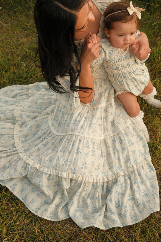 An adult wearing a long, ruffled, floral Ivy City Co dress with a smocked bodice sits on the grass with a baby girl on her lap. Both are dressed in matching light blue outfits, with the baby in the adorable Baby Roselyn Romper in Ivory Porcelain Fleur by Ivy City Co and wearing a headband adorned with a bow.