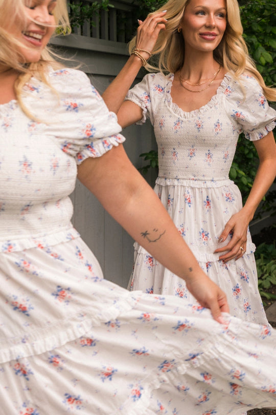 Two women in light Emily Dress in Heritage Soft Ditsy Floral by Ivy City Co smile outdoors. One twirls her skirt, revealing a small star and moon tattoo on her arm. Both wear these nursing-friendly, floral midi dresses that blend style and comfort—perfect for motherhood. Greenery and a wooden fence make up the background.