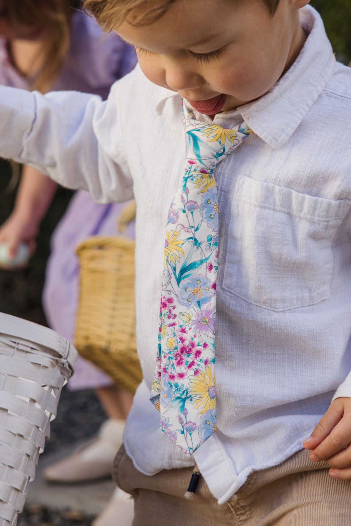 A young child wears the Ivy City Co “Ties in Pretty Plumeria,” a vibrant floral accessory perfect for spring celebrations, paired with a white shirt and holding a white basket. In the background, another child dressed in purple carries a woven basket.