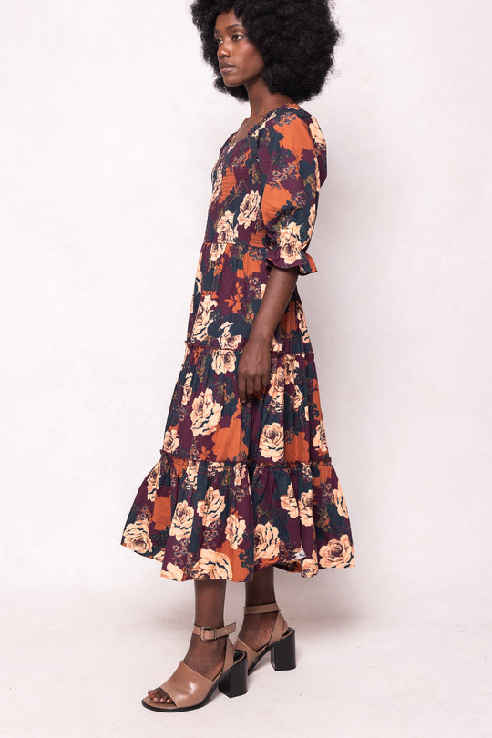 A woman with natural curly hair wears the Ivy City Co Madeline Dress in Equinox Rose, a long, floral-patterned midi dress featuring ruffled sleeves and a tiered, maternity-friendly skirt. She stands sideways in beige heeled sandals against a plain light background.