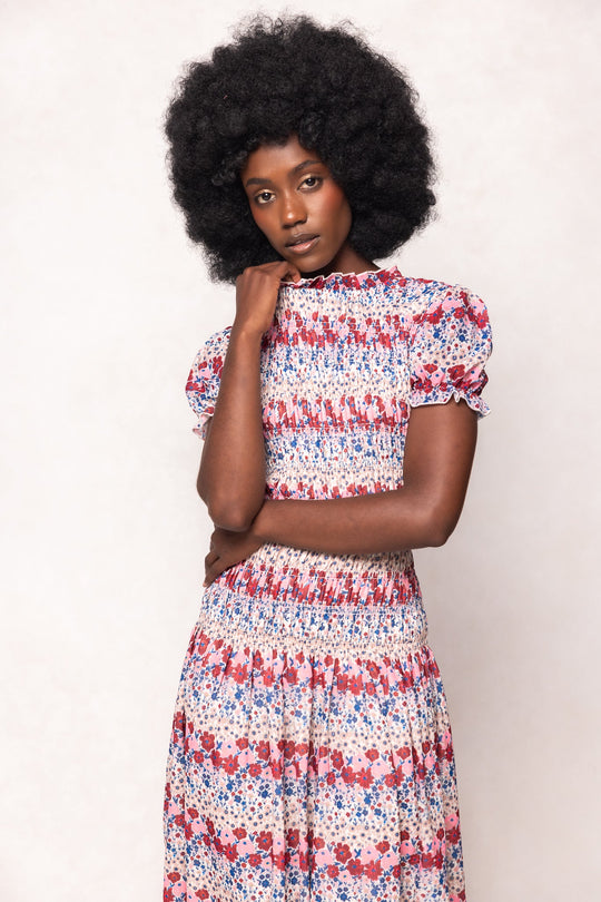 A woman with a natural afro hairstyle poses thoughtfully, wearing the Hayden Dress in Lush Garden by Ivy City Co—a maternity-friendly, short-sleeved floral midi dress showcasing a vibrant red, blue, and white pattern against a pale background.