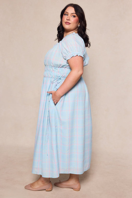 A woman stands sideways wearing the Thalia Dress in Pastel Plaid by Ivy City Co, which features short puff sleeves and practical pockets. Her wavy dark hair and beige flats complement the ensemble as she poses against a neutral background. Maternity-friendly, this dress offers effortless style and comfort, making it an ideal choice for any spring occasion.