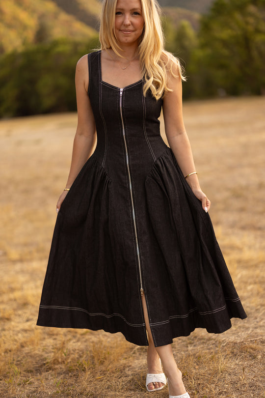 A woman stands outdoors on dry grass, wearing the Ivy City Co Edith Dress in Black—a nursing-friendly, sleeveless tea-length dress with a front zipper—and white sandals. Trees and hills are visible in the blurred background.