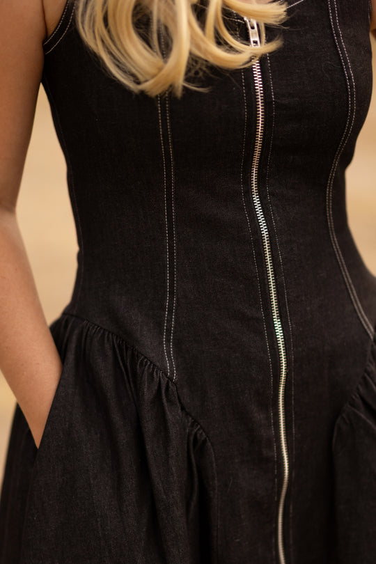 A close-up of a person wearing the Ivy City Co Edith Dress in Black—a sleeveless, tea-length style featuring white stitching and a visible zipper. With their hand tucked into a pocket and blonde hair draped over one shoulder, the image highlights the dress’s nursing-friendly design.