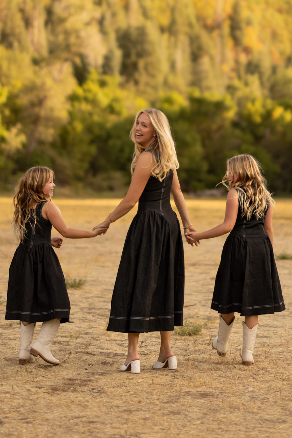 A woman and two young girls wearing matching Ivy City Co Edith Dress in Black—hers is nursing friendly—and white boots hold hands in a dry, grassy field with trees in the background.