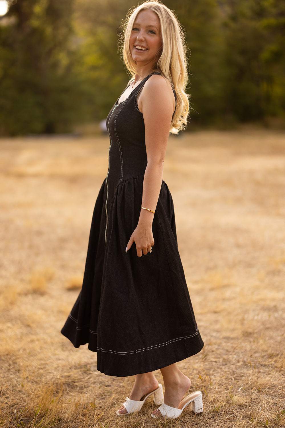 A woman with long blonde hair, wearing the sleeveless Ivy City Co Edith Dress in Black and white heels, stands smiling in a sunlit field of dry grass with trees in the background. This nursing-friendly, tea-length dress from Ivy City Co combines elegant style and comfortable practicality for any occasion.