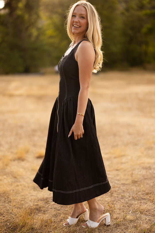 A woman with long blonde hair, wearing the sleeveless Ivy City Co Edith Dress in Black and white heels, stands smiling in a sunlit field of dry grass with trees in the background. This nursing-friendly, tea-length dress from Ivy City Co combines elegant style and comfortable practicality for any occasion.