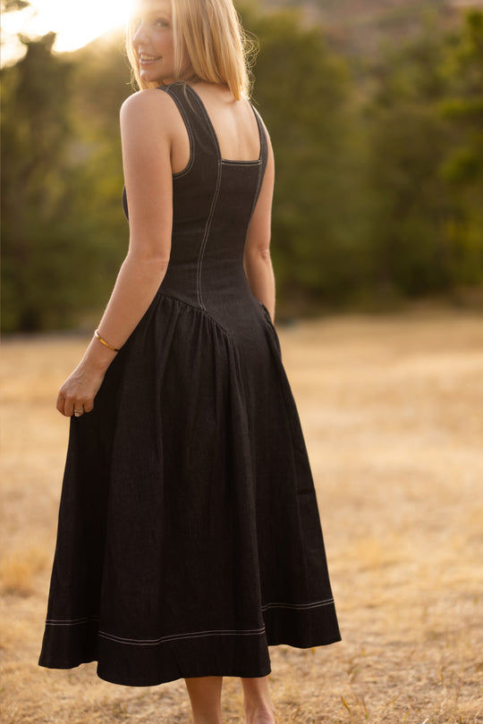 A woman in the sleeveless Edith Dress in Black by Ivy City Co stands in a field of dry grass, gazing over her shoulder. Sunlight shines behind her, casting a warm, glowing effect, while trees appear softly blurred in the background.
