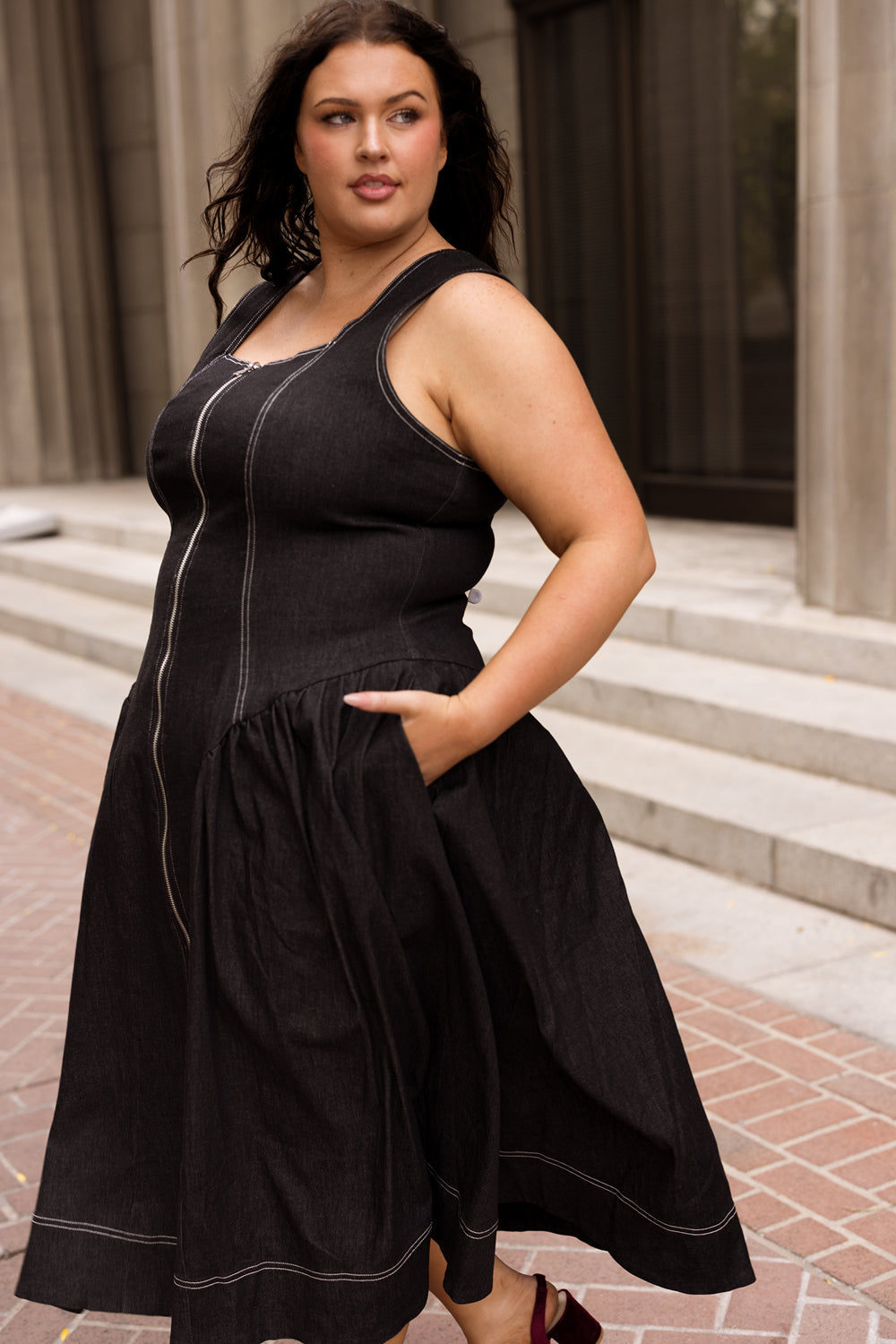 A woman wearing the Edith Dress in Black by Ivy City Co—a sleeveless, tea-length dress with white stitching—stands confidently on a stone walkway before an elegant columned building, one hand in her pocket. This nursing-friendly silhouette from Ivy City Co effortlessly combines elegance and practicality.