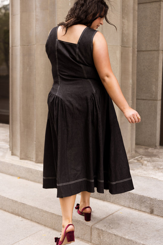 Wearing the Ivy City Co Edith Dress in Black—an elegant, nursing-friendly tea-length style accented with white stitching—a woman in red velvet heels walks up stone steps beside towering columns.