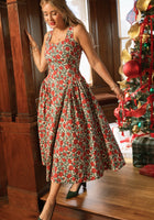 A woman stands indoors near a decorated Christmas tree with presents beneath it, smiling as she wears the Ivy City Co Edith Dress in Red Roses paired with green shoes.