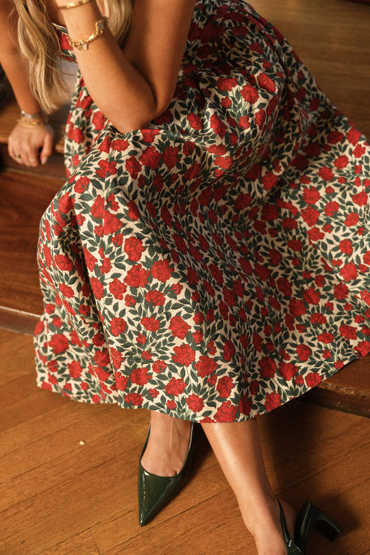 A woman sits with her legs crossed on a wooden floor, wearing the Ivy City Co Edith Dress in Red Roses—a sleeveless dress featuring a red rose and green leaf print—paired with dark green pointed heels. Her face is not visible.