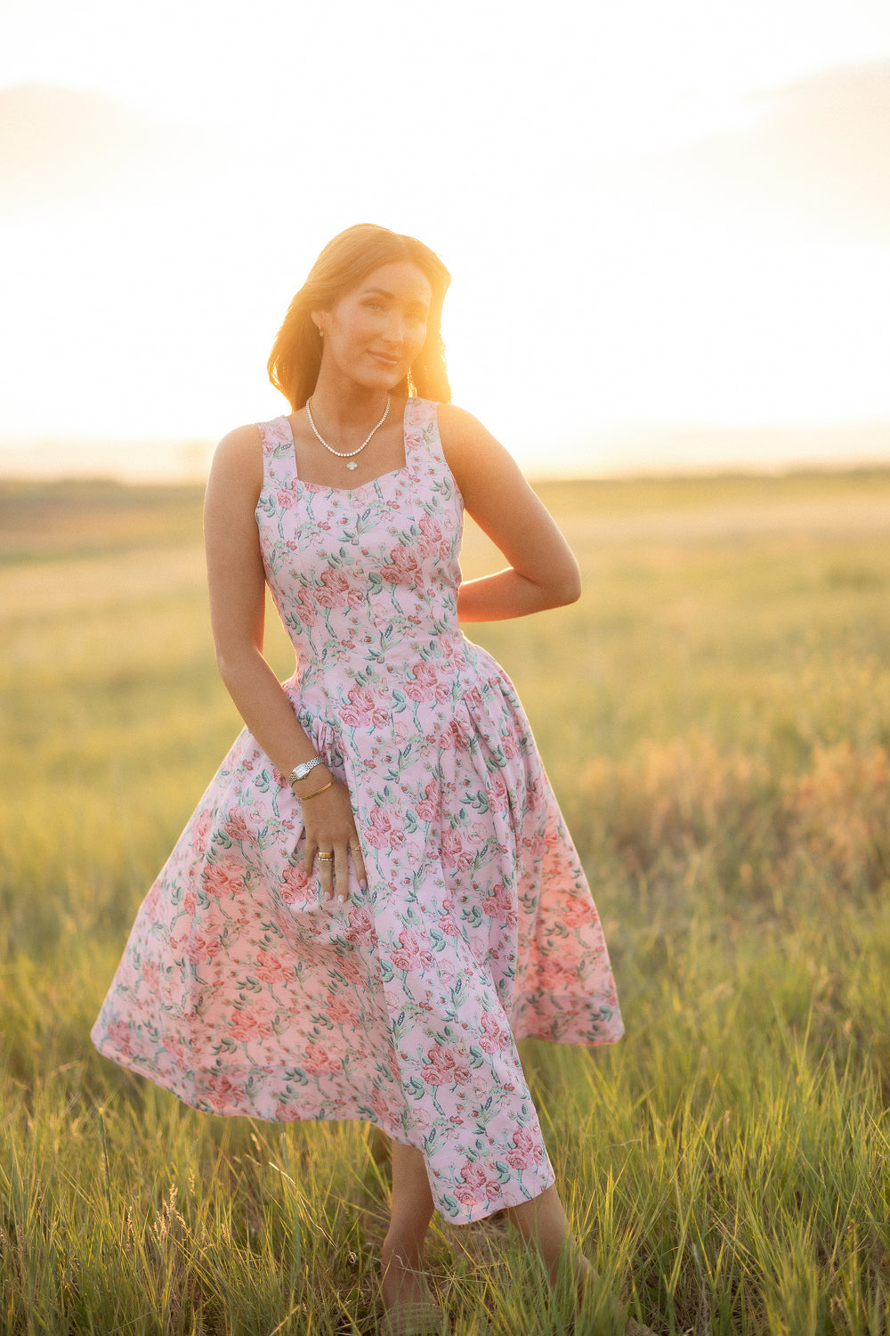 A woman wearing the Edith Dress in Pink Sketched Flower by Ivy City Co stands in a sunlit field, smiling gently as she holds her dress. The soft sunlight creates a warm, glowing effect around her.