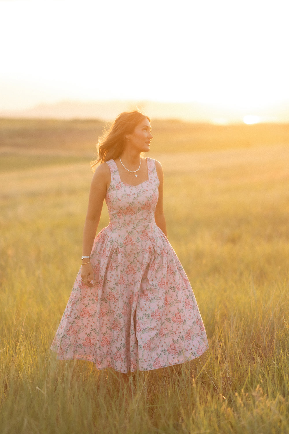 A woman wears the Ivy City Co "Edith Dress in Pink Sketched Flower" as she stands in a sunlit field of tall grass at sunset, looking to the side with a gentle smile as warm light surrounds her.