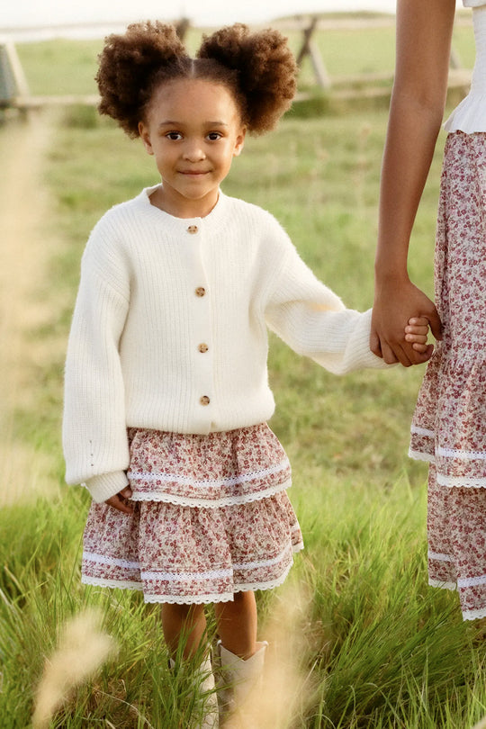 A young girl with curly hair, dressed in a white sweater and the Ivy City Co Mini Annalise Skirt in Philocaly Floral—a girls’ cotton skirt featuring a delicate floral pattern—stands on grass holding an adult’s hand.