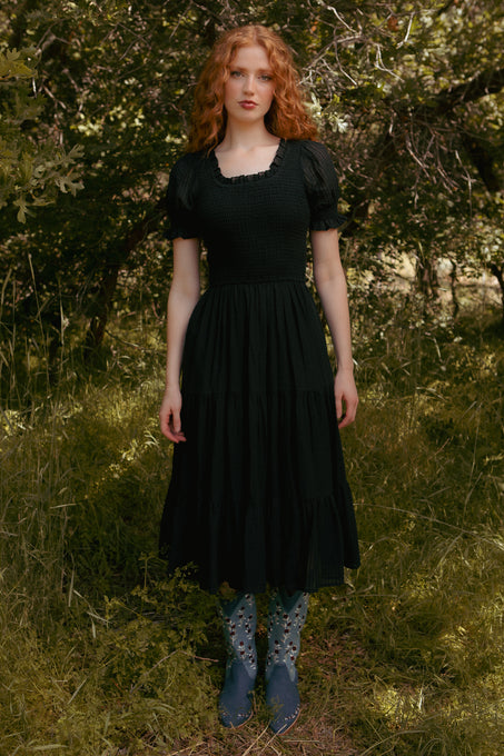 A woman with long red hair stands in a sunlit forest wearing the Ivy City Co Ellery Dress in Black, paired with blue floral boots. She looks at the camera, surrounded by greenery; her stylish outfit is also maternity friendly.
