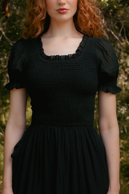 A woman with curly red hair stands outdoors among green foliage, wearing the Ellery Dress in Black by Ivy City Co. The focus is on her upper body, her face partially visible, and the dresss smocked cotton detail is highlighted.
