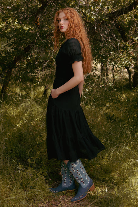 A person with long curly red hair stands sideways in a sunlit, wooded area wearing the Ivy City Co Ellery Dress in Black, a maternity-friendly midi, paired with blue cowboy boots decorated with white stars.