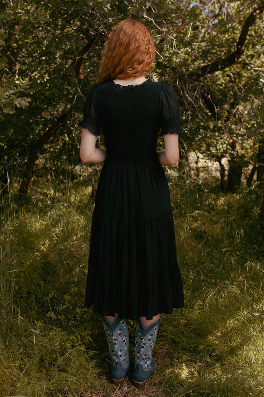 A person with long curly red hair stands in a sunlit grassy forest, facing away, wearing the Ivy City Co Ellery Dress in Black and light blue floral cowboy boots; the look is stylish and maternity-friendly.