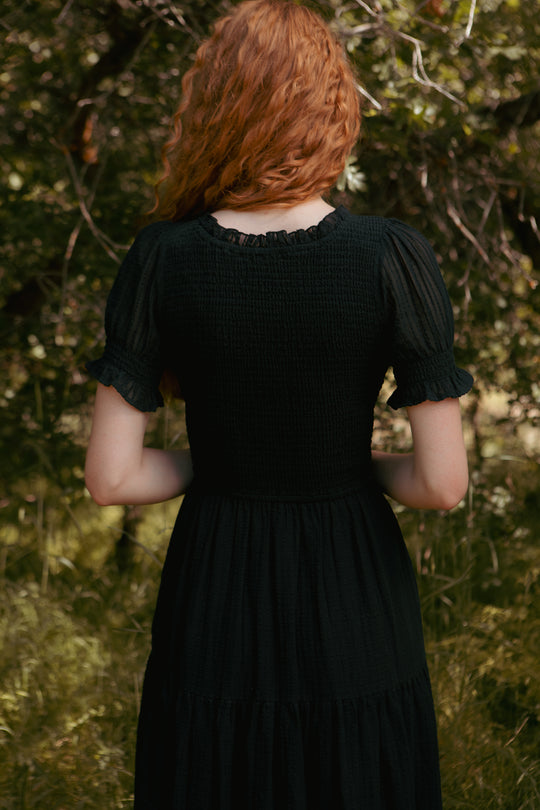 A person with long, wavy red hair stands outdoors among lush greenery, wearing the Ellery Dress in Black by Ivy City Co—an effortless blend of style and natural beauty.