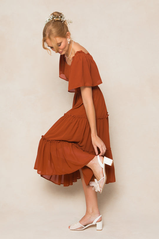 A woman wearing the Ellie Dress in Amber Chiffon by Ivy City Co bends down to adjust her white heeled sandal. Flowers adorn her hair as she stands against a neutral background.