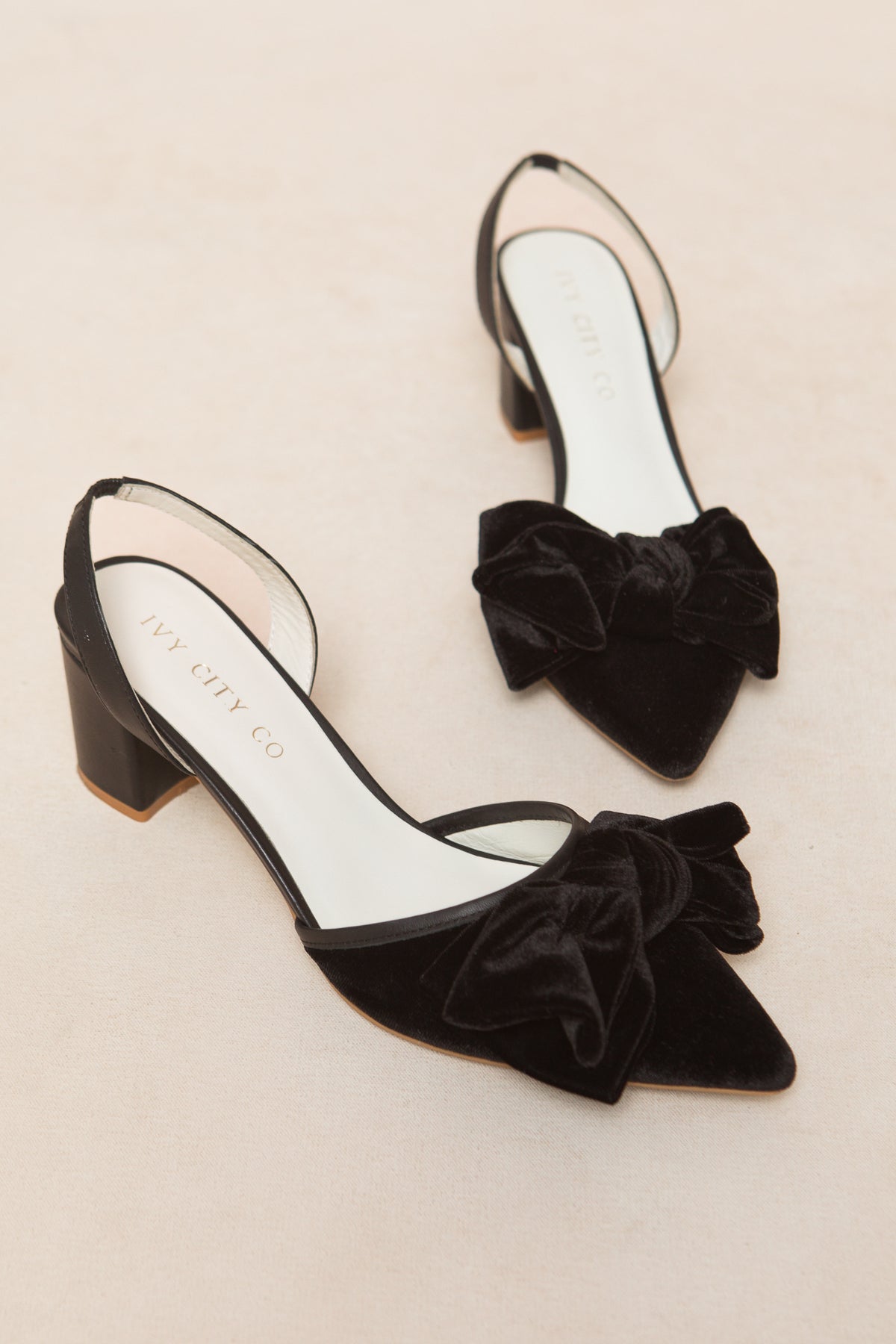 The Eloise Bow Heel in Black by Ivy City Co features black velvet slingbacks with pointed toes, low block heels, large velvet bows on the toes, and a white branded insole, displayed on a light beige surface.