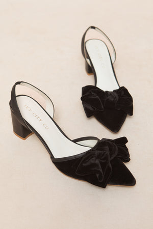 The Eloise Black Bow Heel by Ivy City Co features black velvet, large front bows, a pointed toe, and a low block heel. Displayed on a light beige surface, its branded insole adds an elegant touch to these chic heels.