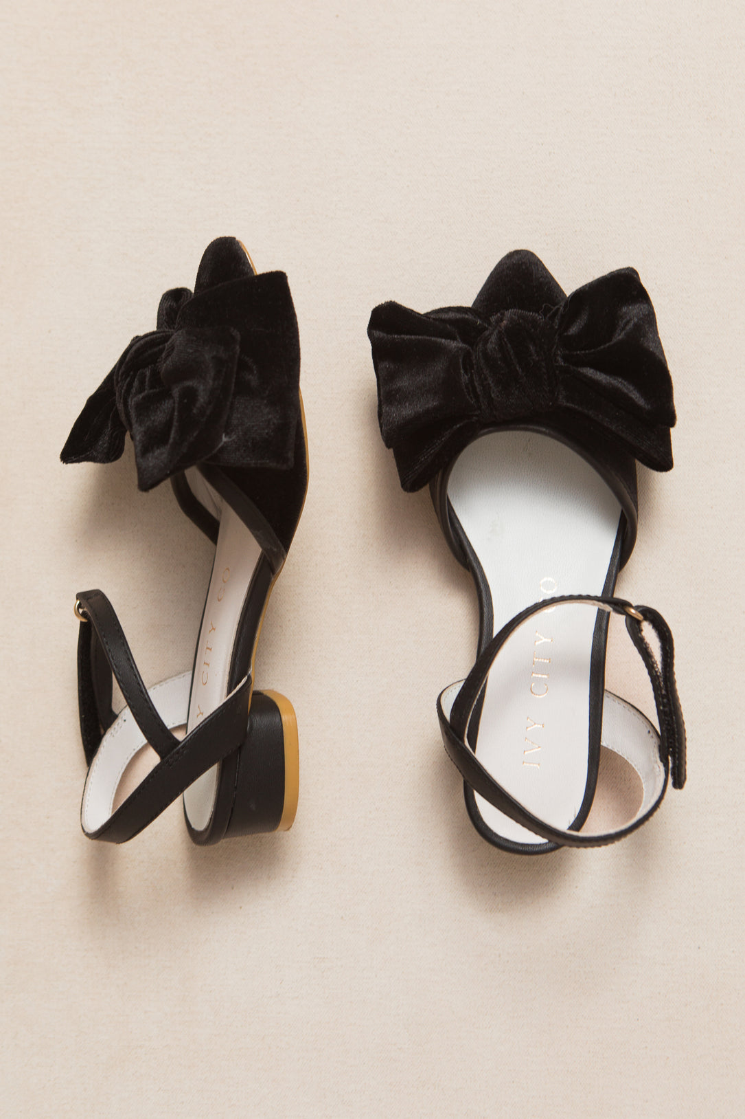 The Ivy City Co Mini Eloise Black Bow Heel sandals—black, open-toe, with ankle straps and large velvet bows—are displayed on a light beige surface.