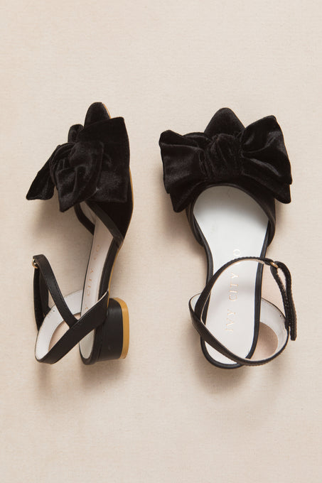 The Ivy City Co Mini Eloise Black Bow Heel sandals—black, open-toe, with ankle straps and large velvet bows—are displayed on a light beige surface.