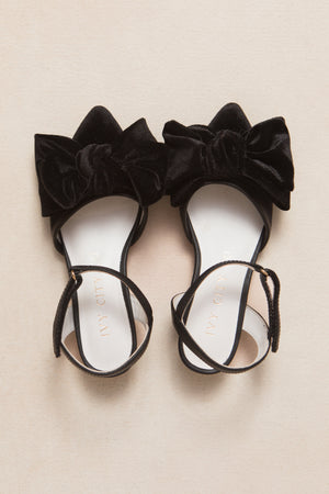 The Mini Eloise Black Bow Heel by Ivy City Co is a black velvet shoe with large front bows and ankle straps, shown on a light beige surface.